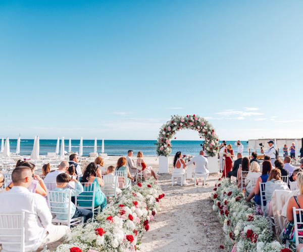 Beach wedding ceremony with floral arch and seated guests.