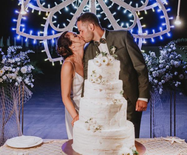 Bride and groom kiss by wedding cake, colorful lights in background.