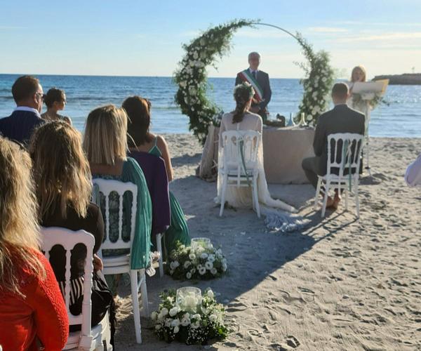 Beach wedding ceremony with floral arch and seated guests.